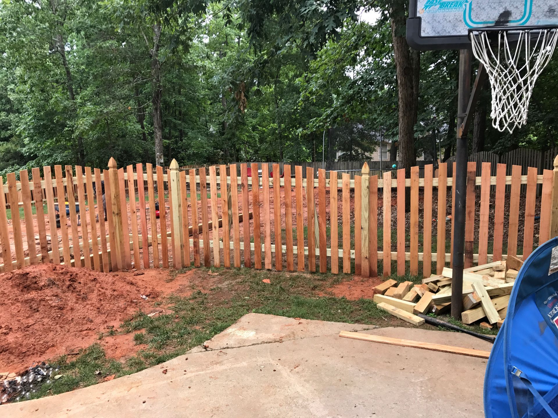 A wooden picket fence being built in a backyard next to a basketball hoop.