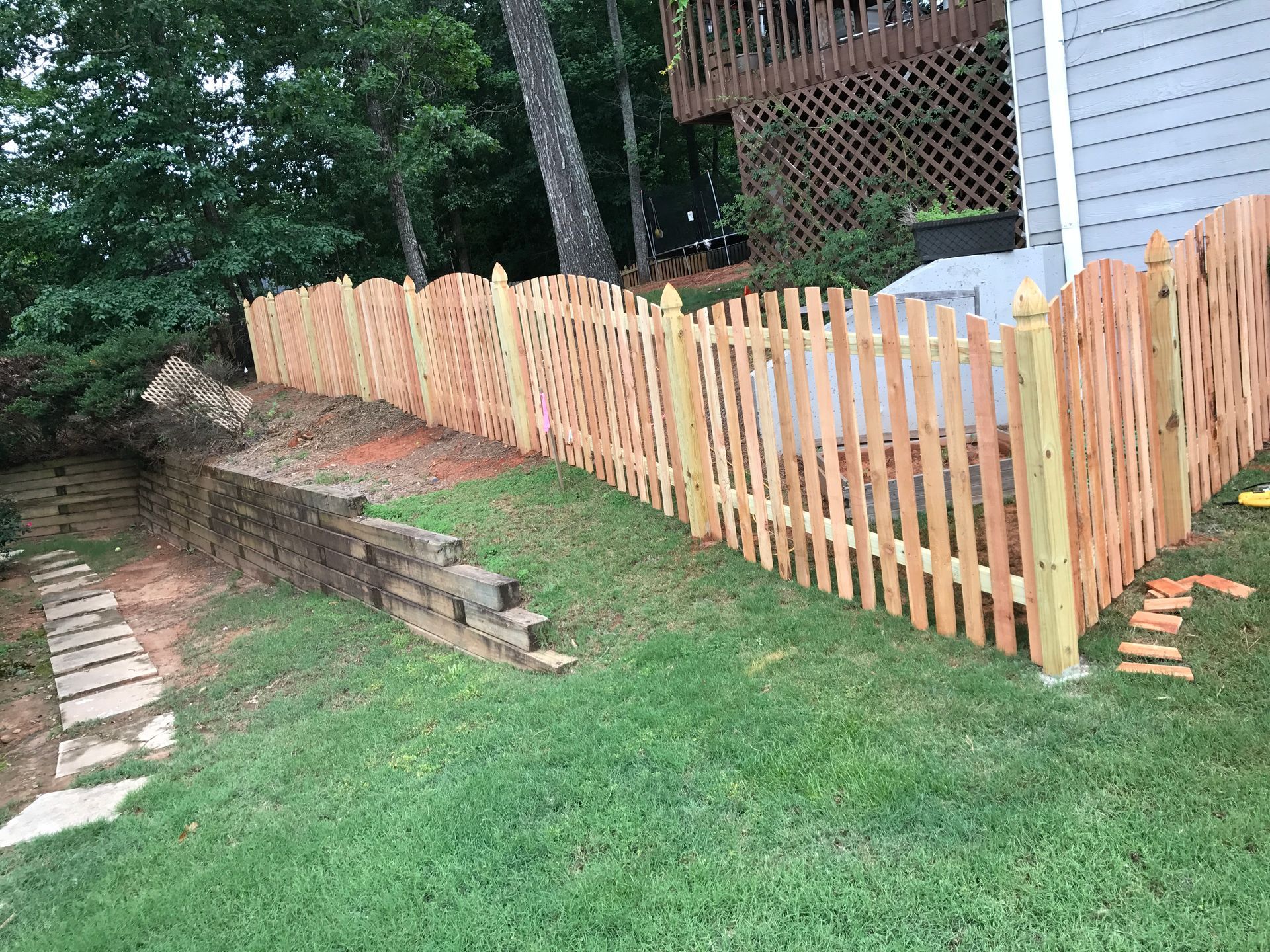 Wooden picket fence on a grassy hill, with a retaining wall and trees in the background.