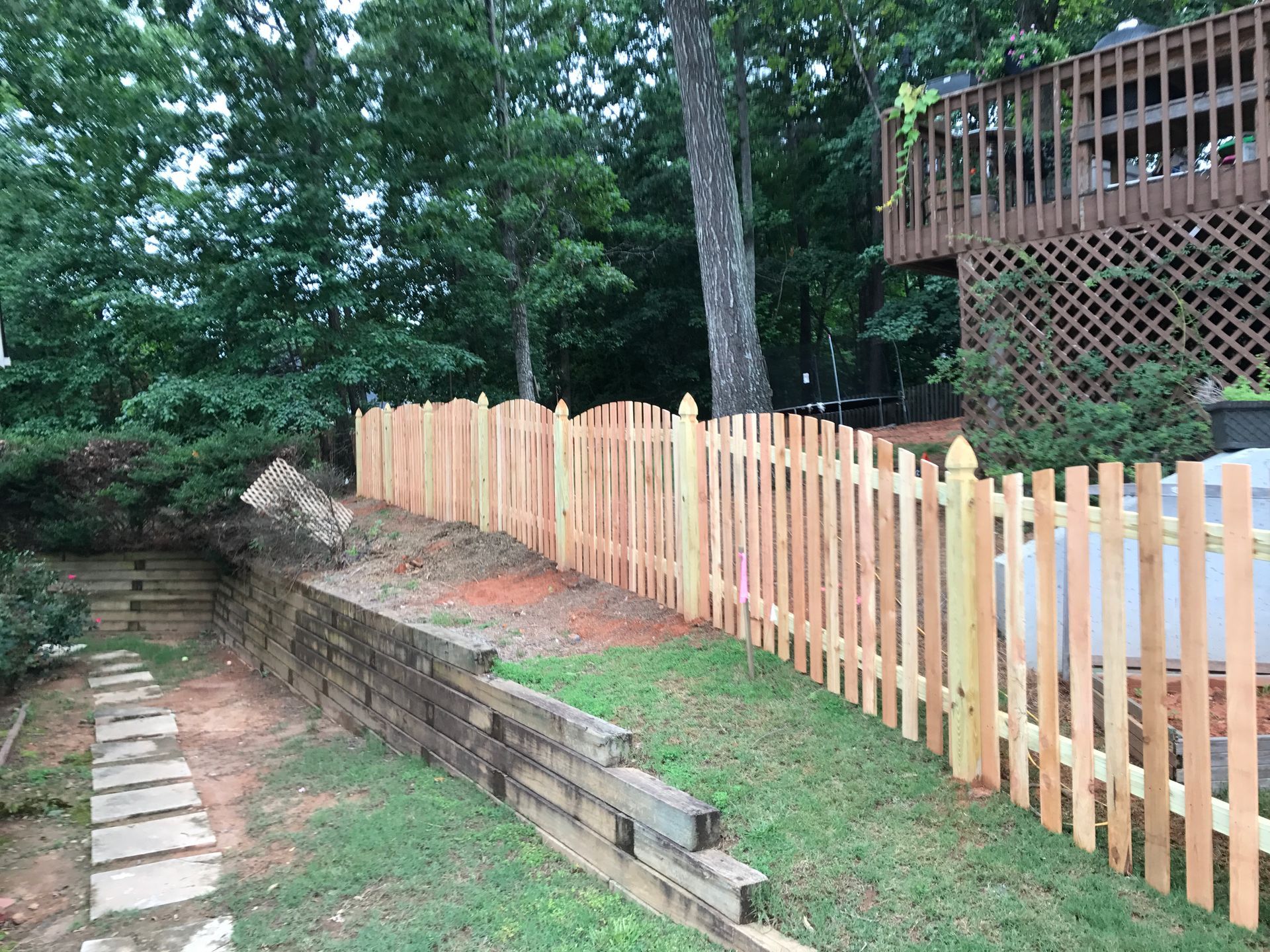 Wooden picket fence in a yard next to a retaining wall and trees.