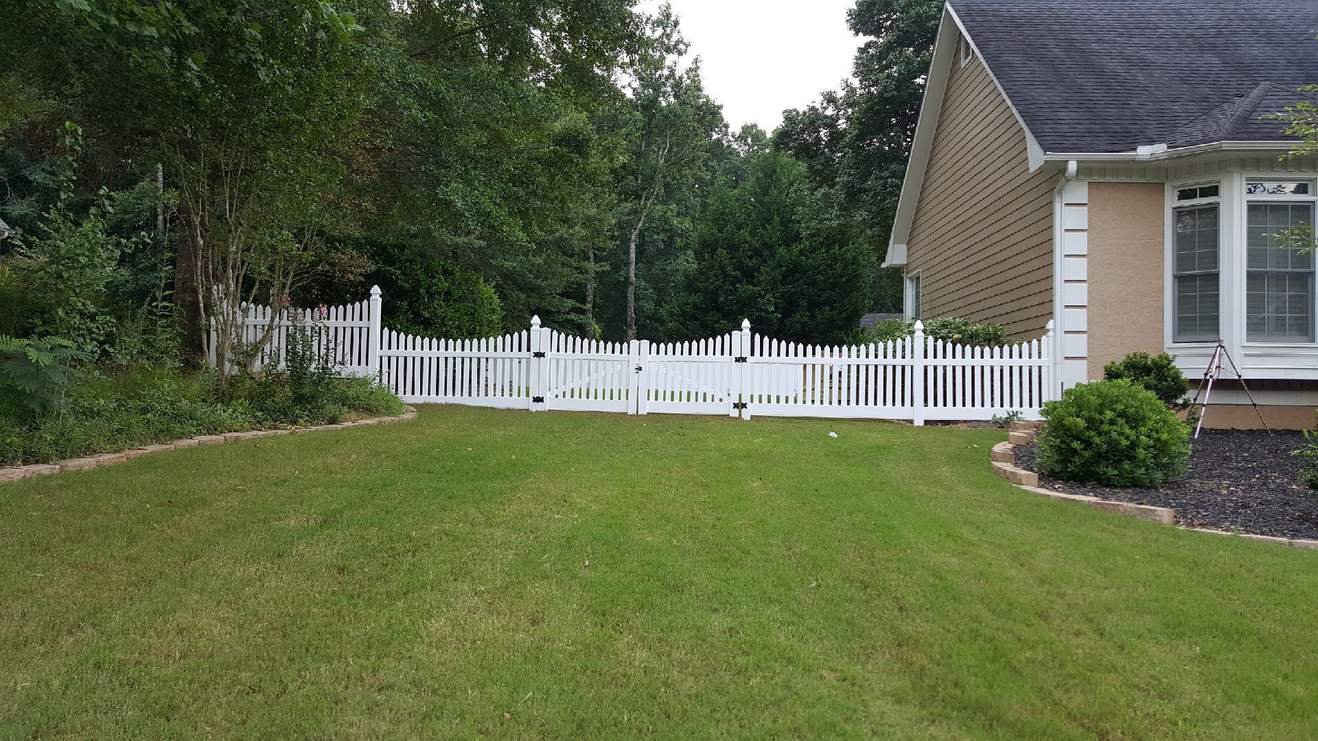 White picket fence in front of a house, separating the green lawn from trees and landscaping.