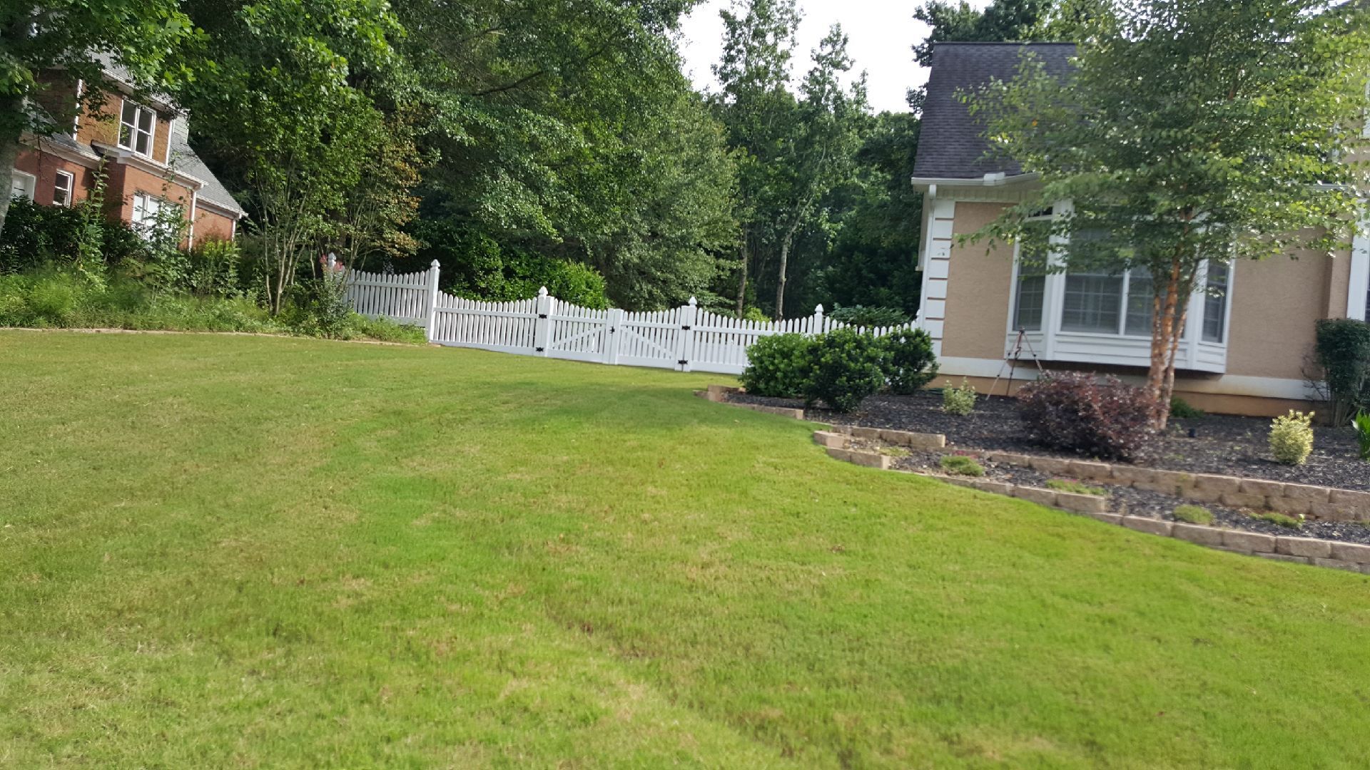 Green lawn slopes toward a white fence and a tan house with a small tree.