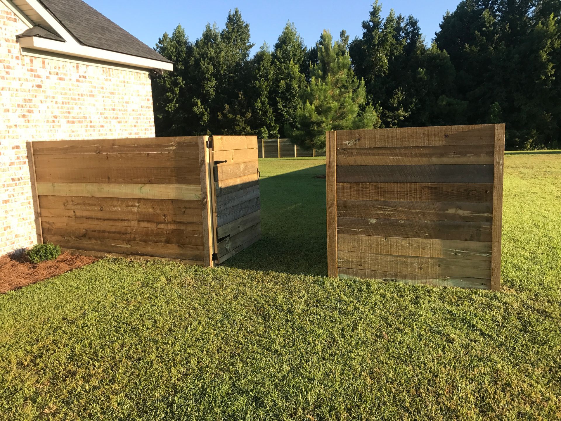 Wooden fence sections frame an open area in a grassy yard, with a brick house and trees in the background.