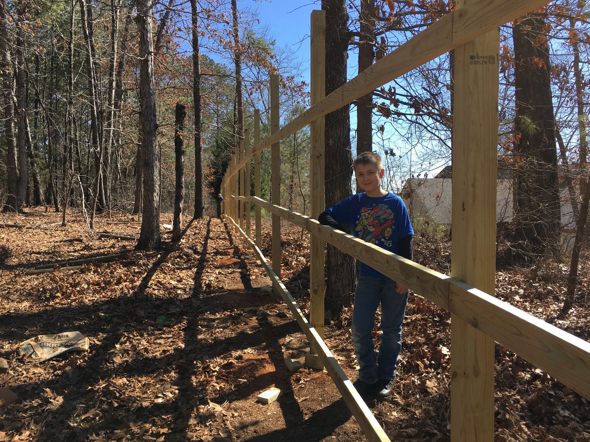 Boy standing beside a new wooden fence in a wooded area on a sunny day.