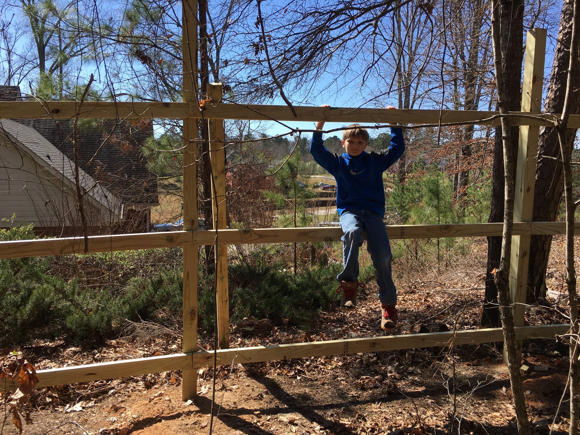 Boy hanging on a wooden fence outdoors, trees and house in the background. Sunny day.
