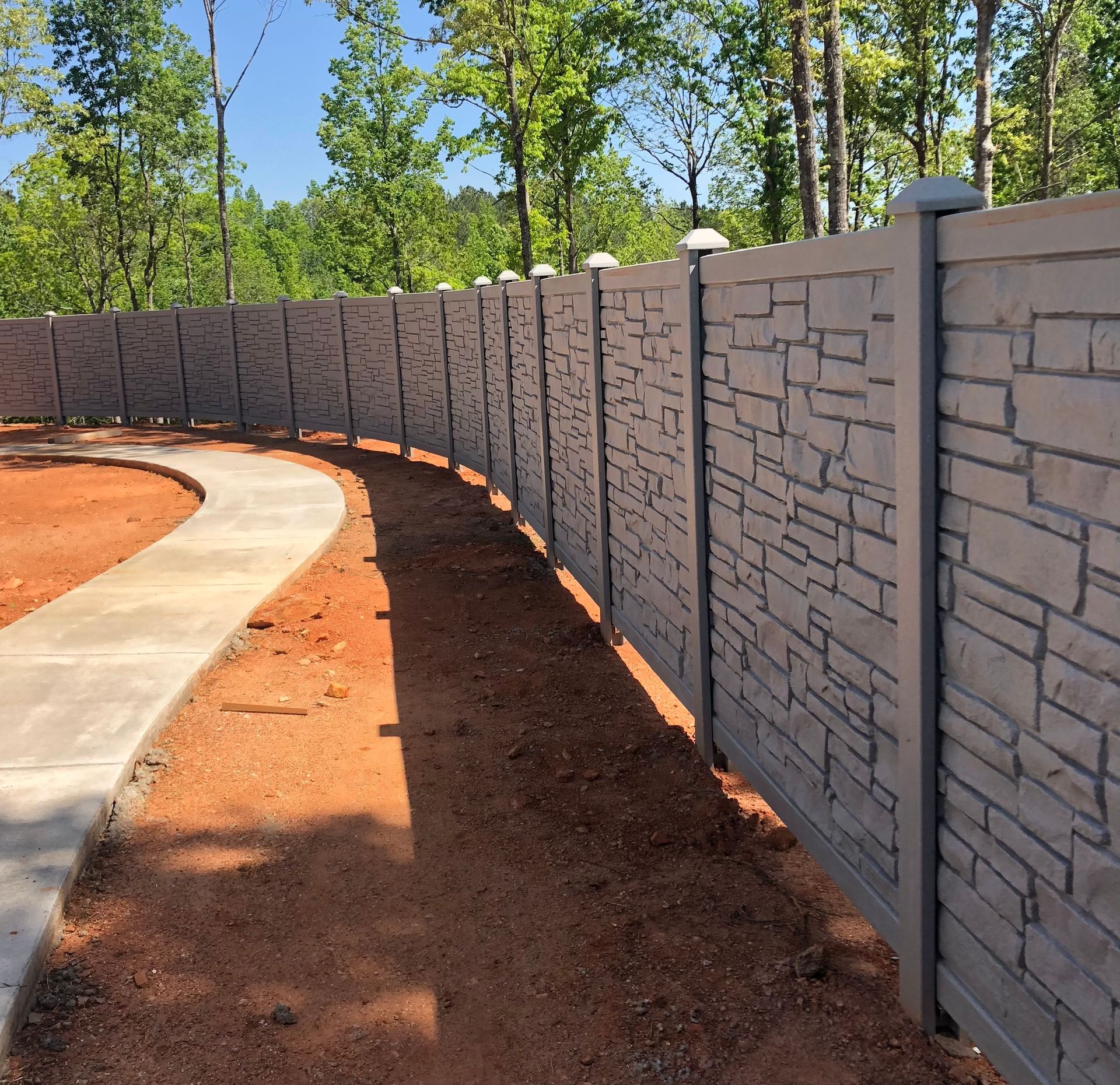Curved gray stone fence with posts, next to a concrete path and red dirt.