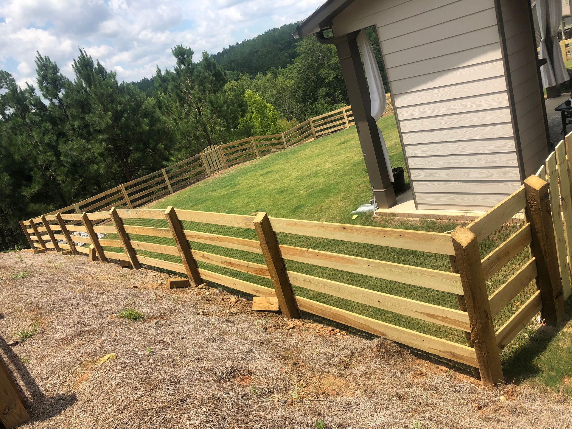 Wooden fence on a hillside, with a house in the background and trees.