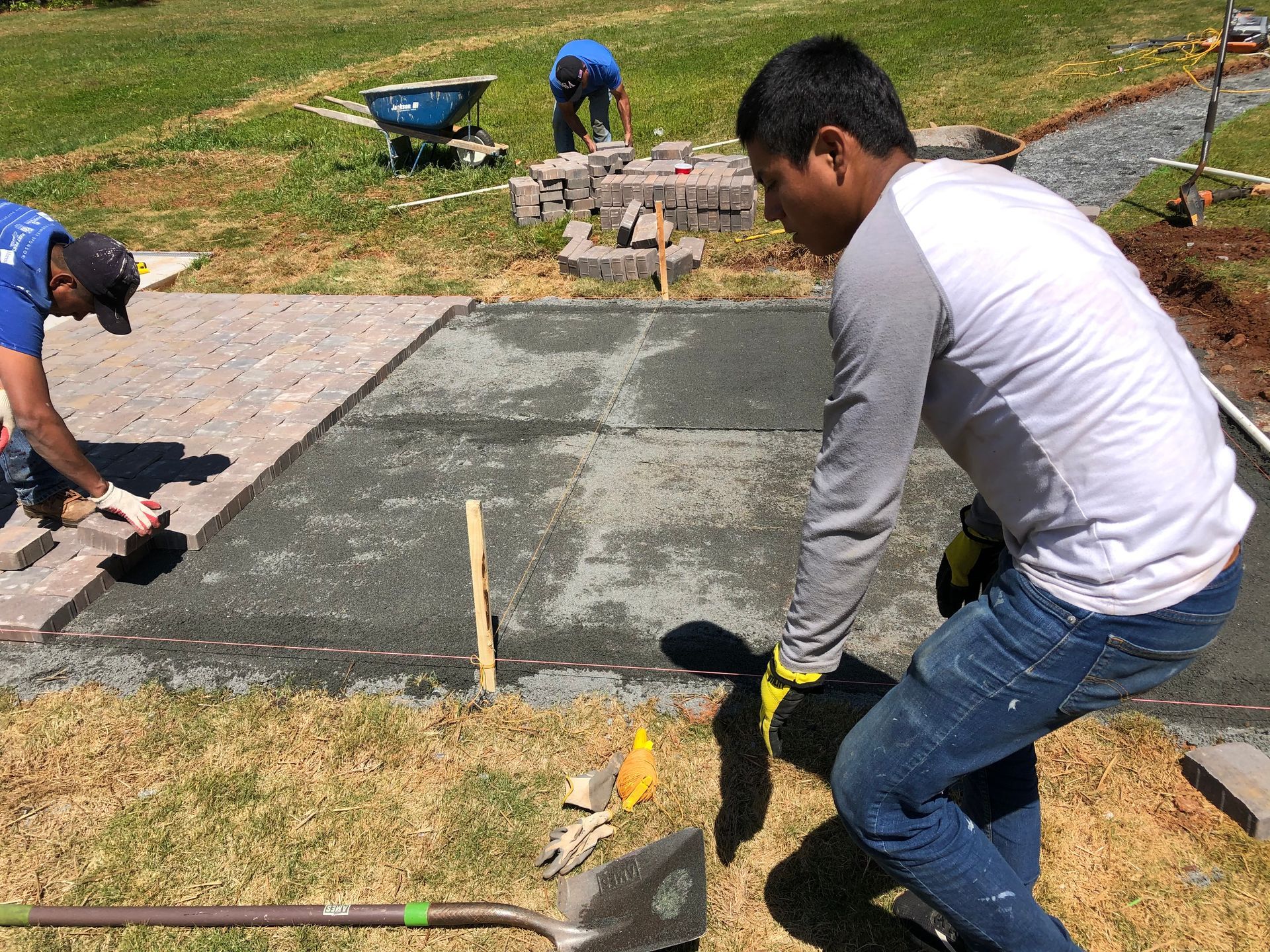 Workers laying paving stones on a patio in a grassy yard; gray and tan colors.