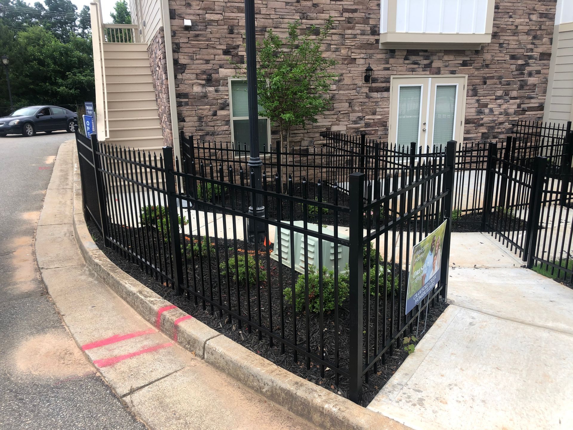 Black metal fence encloses a small planted area next to a building with stone facade.