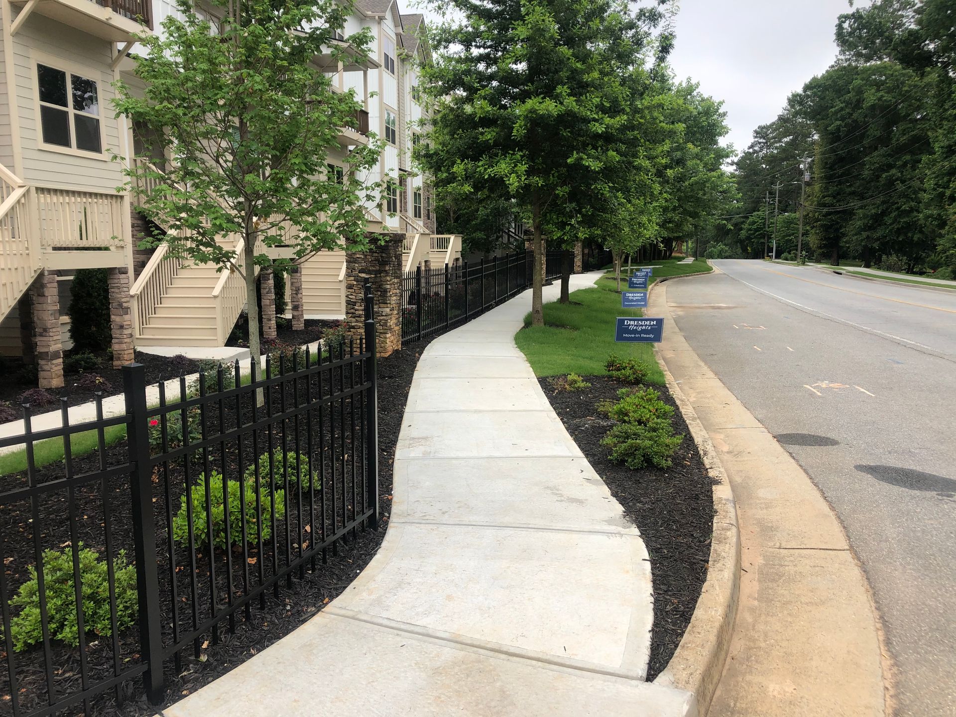 Sidewalk next to black fence, building, trees, and road.