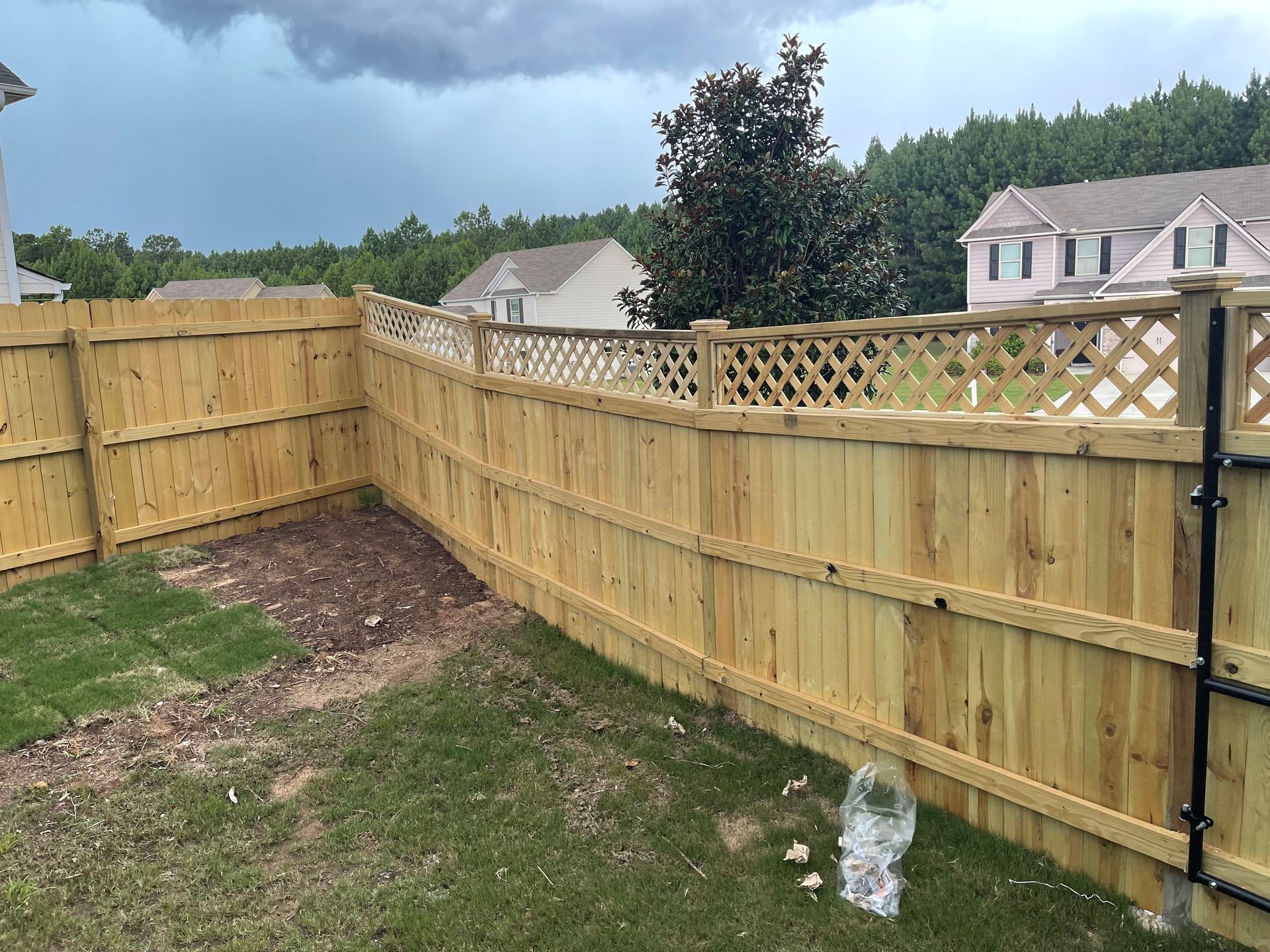 Wooden fence surrounding a grassy yard with houses and trees in the background under a cloudy sky.