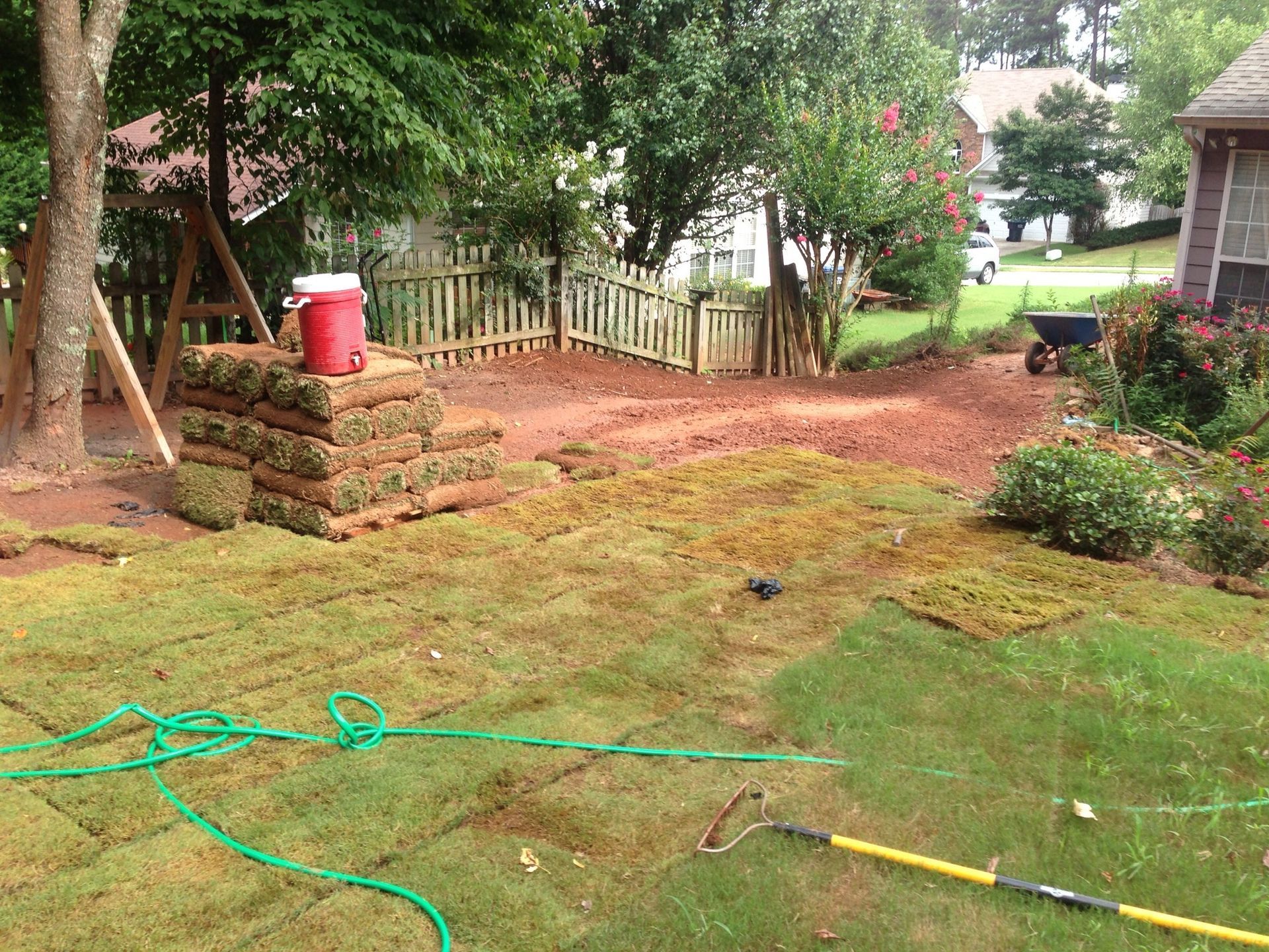Backyard with stacked sod, green grass, red dirt, hose, and a fence.