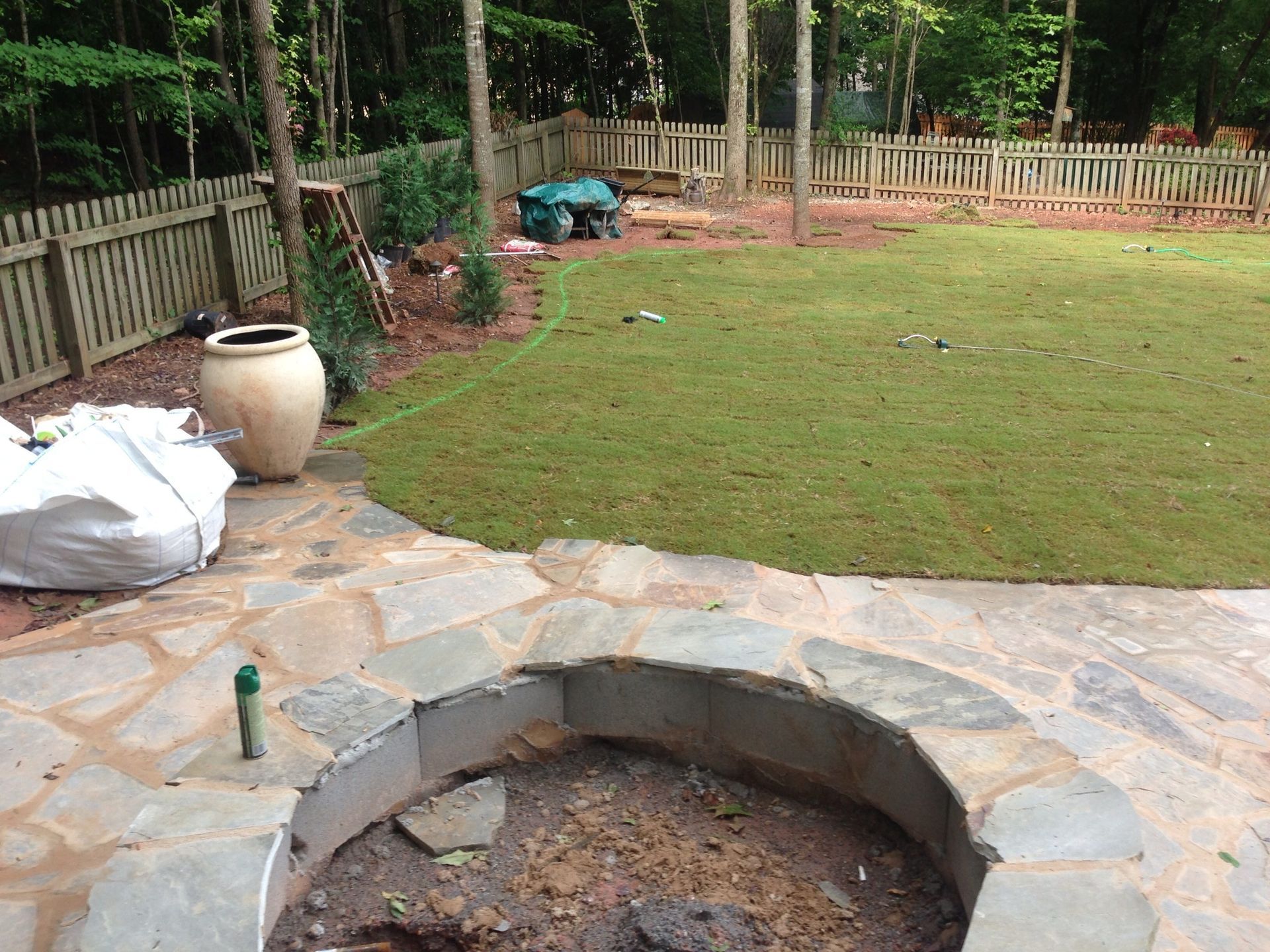 Stone patio with fire pit, looking onto a grassy backyard bordered by a wooden fence and trees.