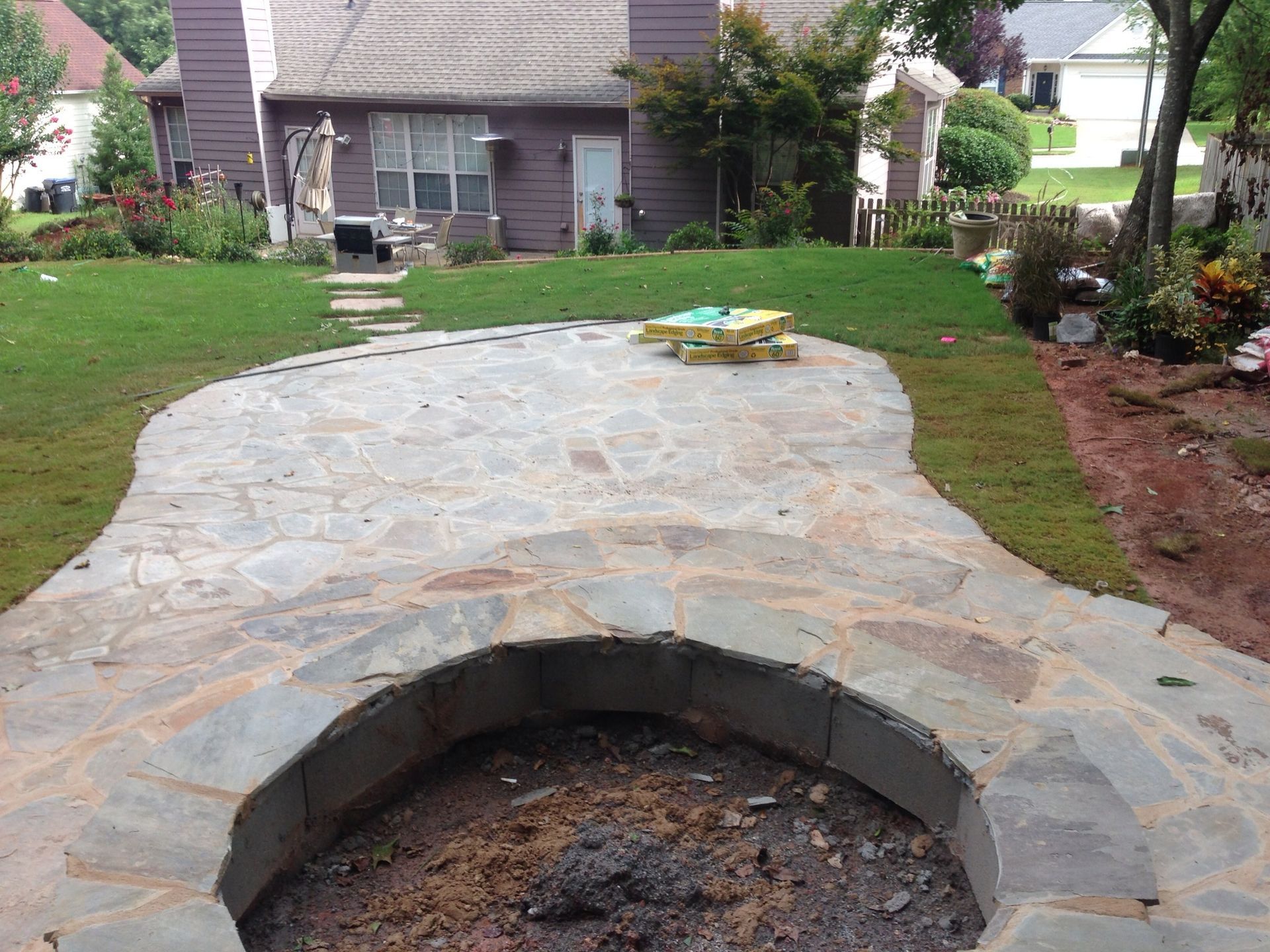 Stone patio with built-in fire pit in a backyard with a house, lawn, and landscaping.
