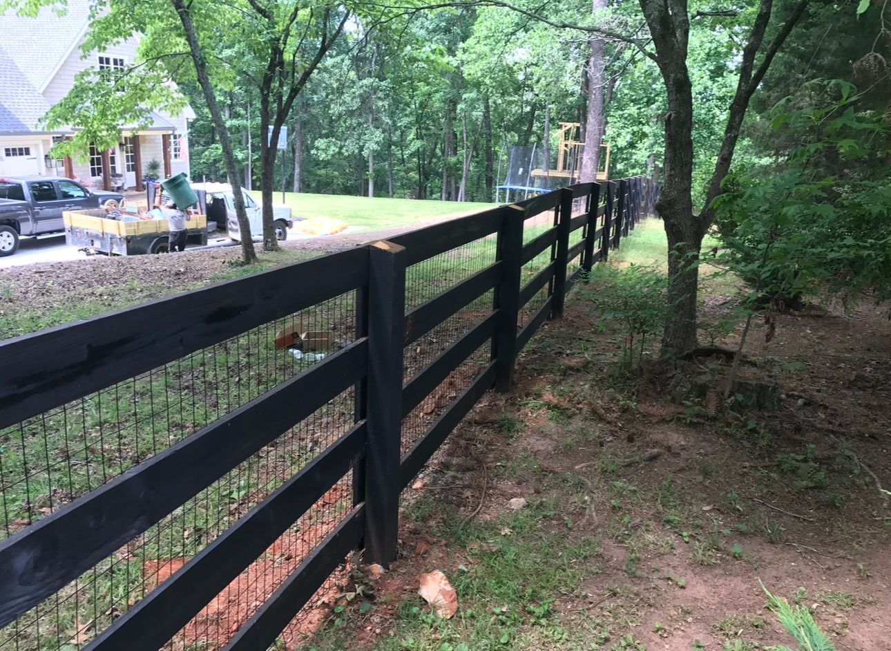 Black wooden fence with wire mesh in a grassy yard, trees in background, house visible.