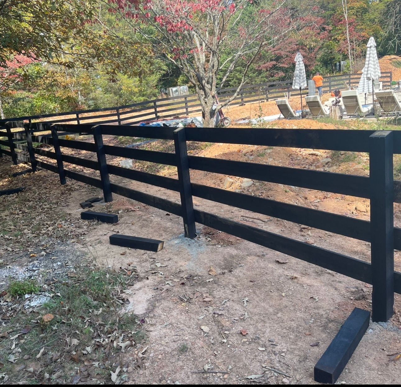 Black wooden fence in a yard with a pool area in the background. Autumn leaves and trees visible.