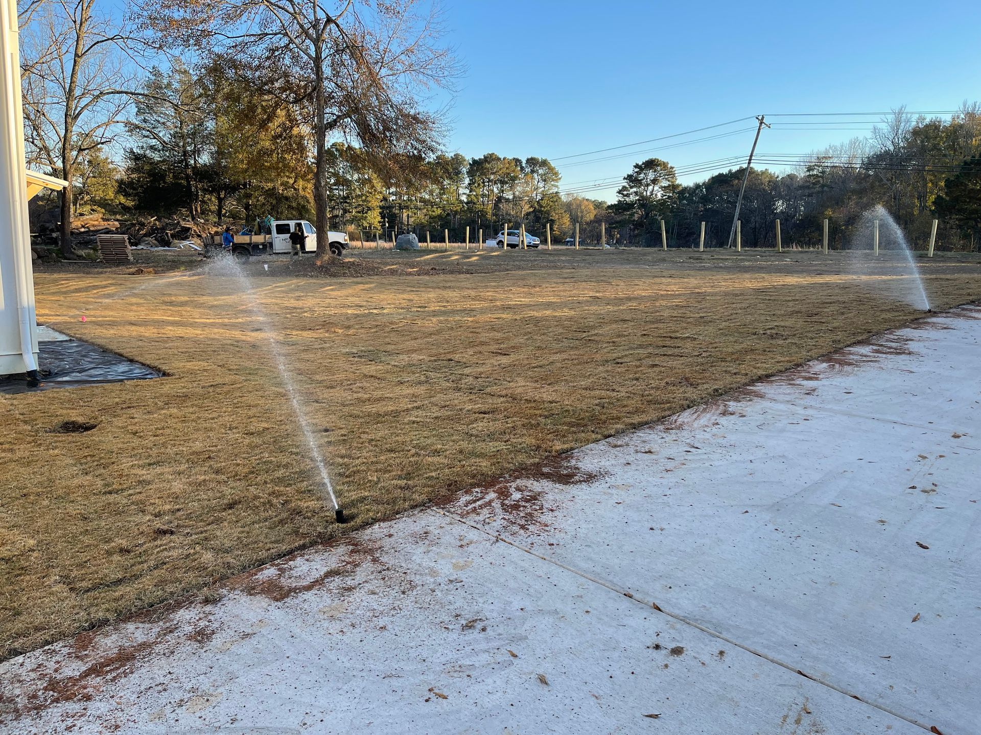 Sprinklers watering a brown, dry field next to a concrete walkway. Trees and blue sky in background.