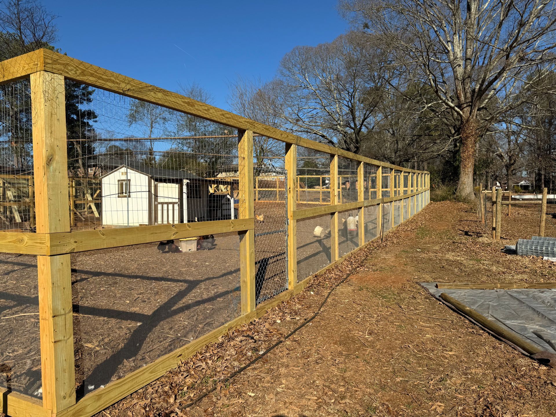 Wooden fence enclosing a dirt area with a small white shed in the background under a blue sky.