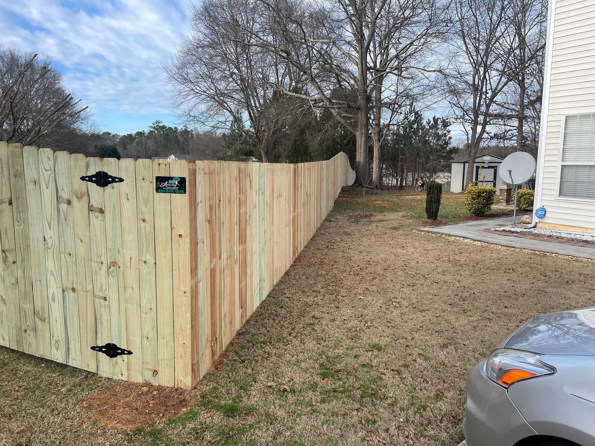 Wooden fence surrounds a yard with a car parked nearby on a brown grassy lawn.