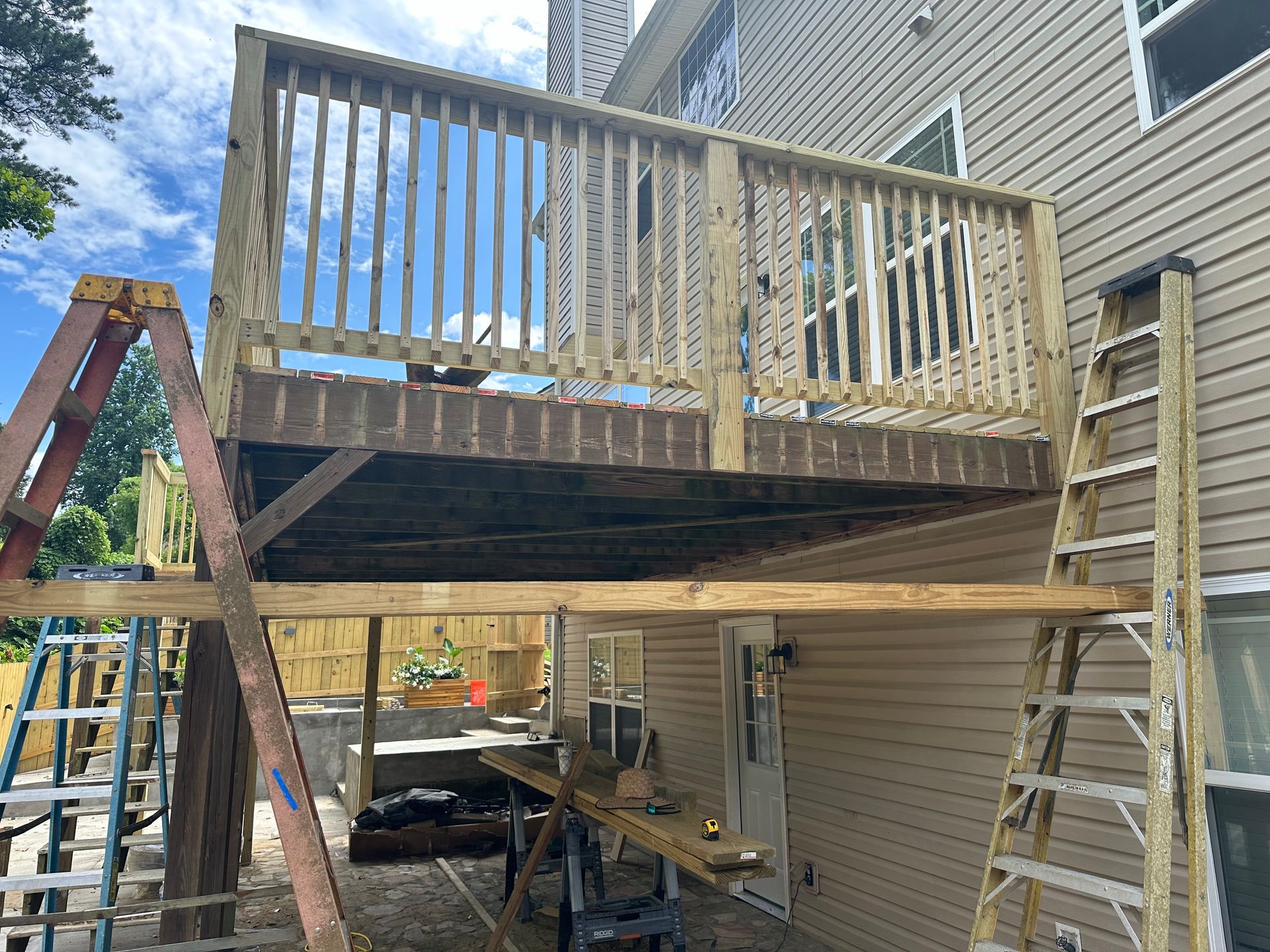 Two-story wooden deck under construction; ladders and tools visible. House siding in the background.