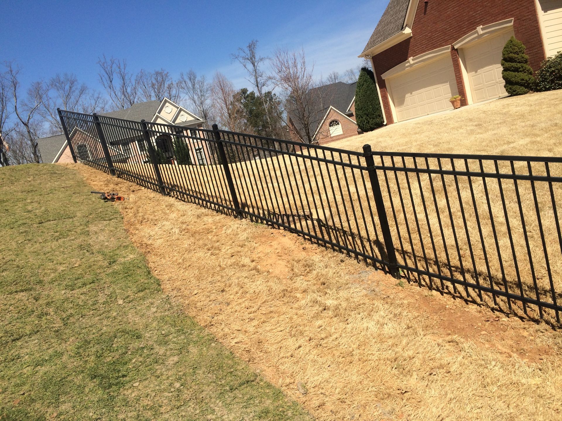 Black metal fence on a grassy hill, separating two yards. A house is in the background.