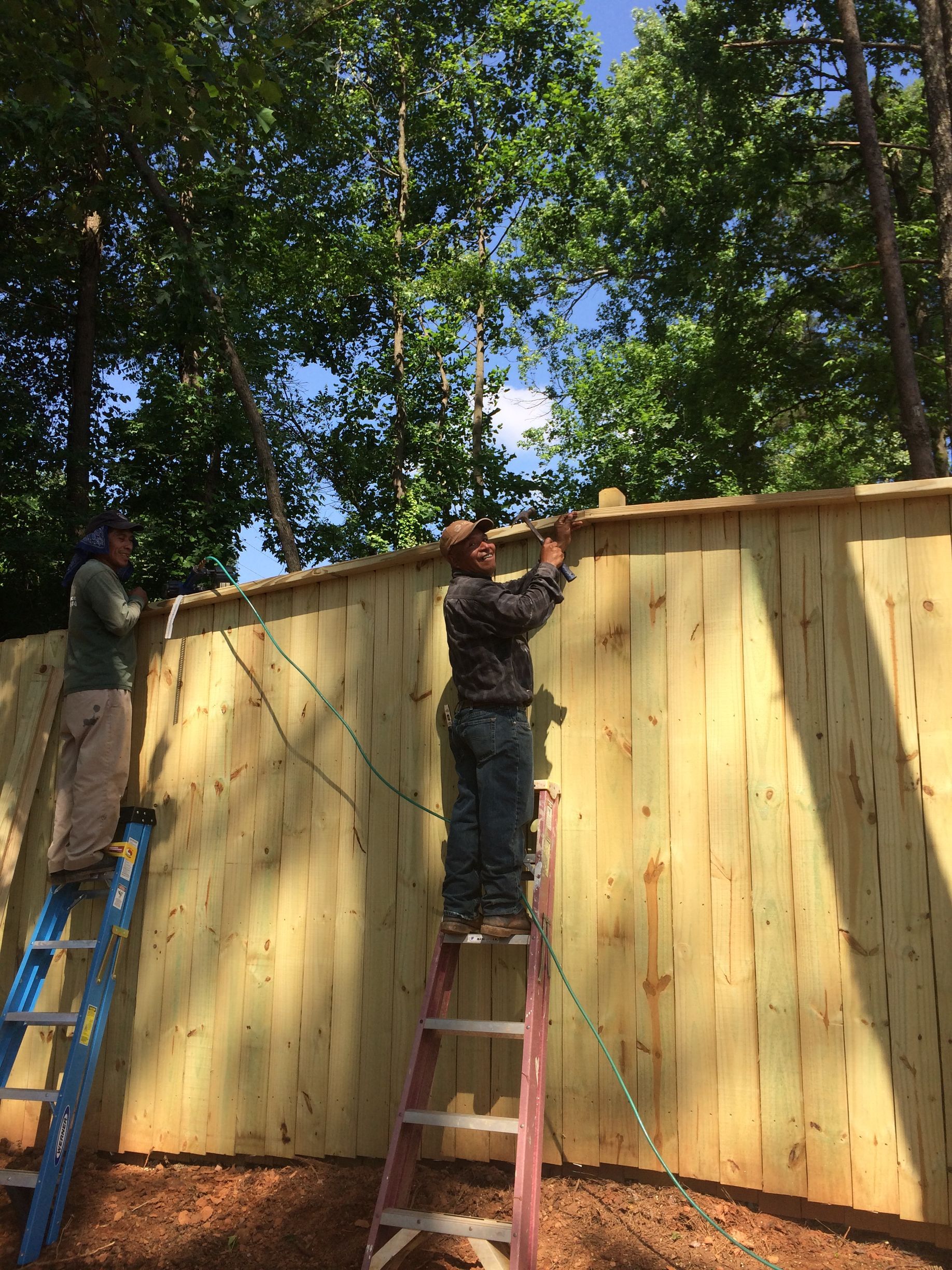 Two people building a wooden fence outdoors on a sunny day.