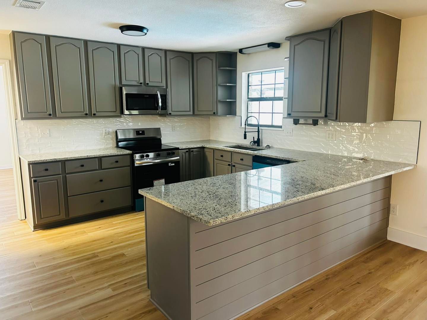 Gray kitchen cabinets and island with granite countertops, stainless steel appliances, and wood floors.