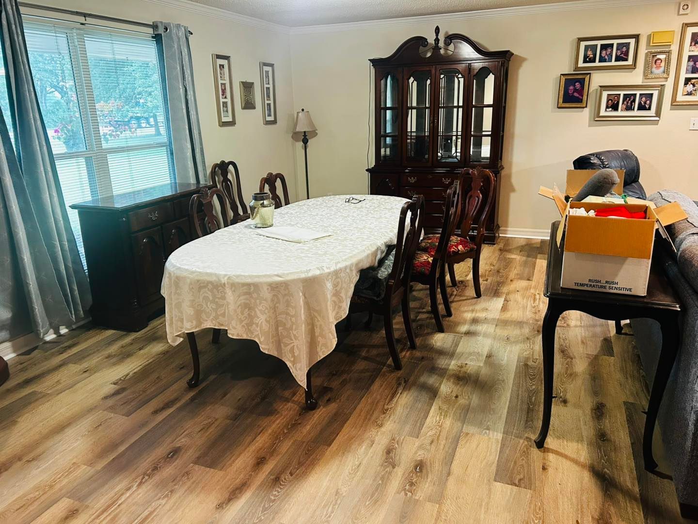Dining room with oval table, hutch, buffet, chairs, and framed photos. Wooden floor, lace tablecloth.