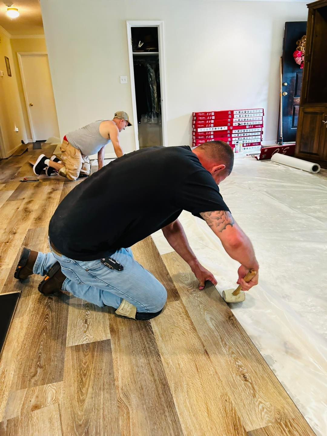 Two people installing wood-look flooring indoors. One kneels, applying adhesive, while the other works in the background.