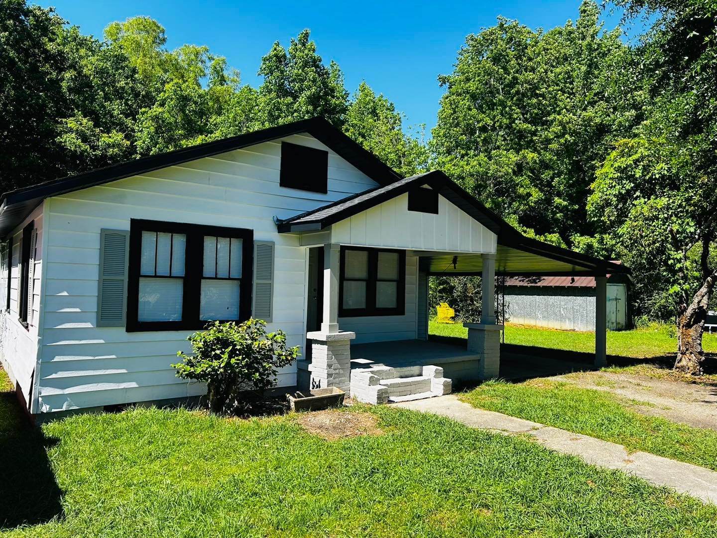 White house with black trim, gray porch, green yard, and trees against a blue sky.