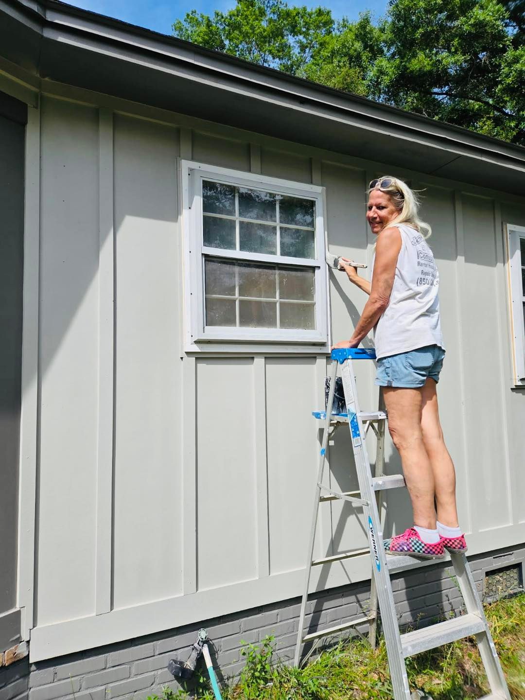 Woman on a ladder painting a house exterior, smiling. Wearing shorts, t-shirt, and Crocs. Light gray siding and white trim.