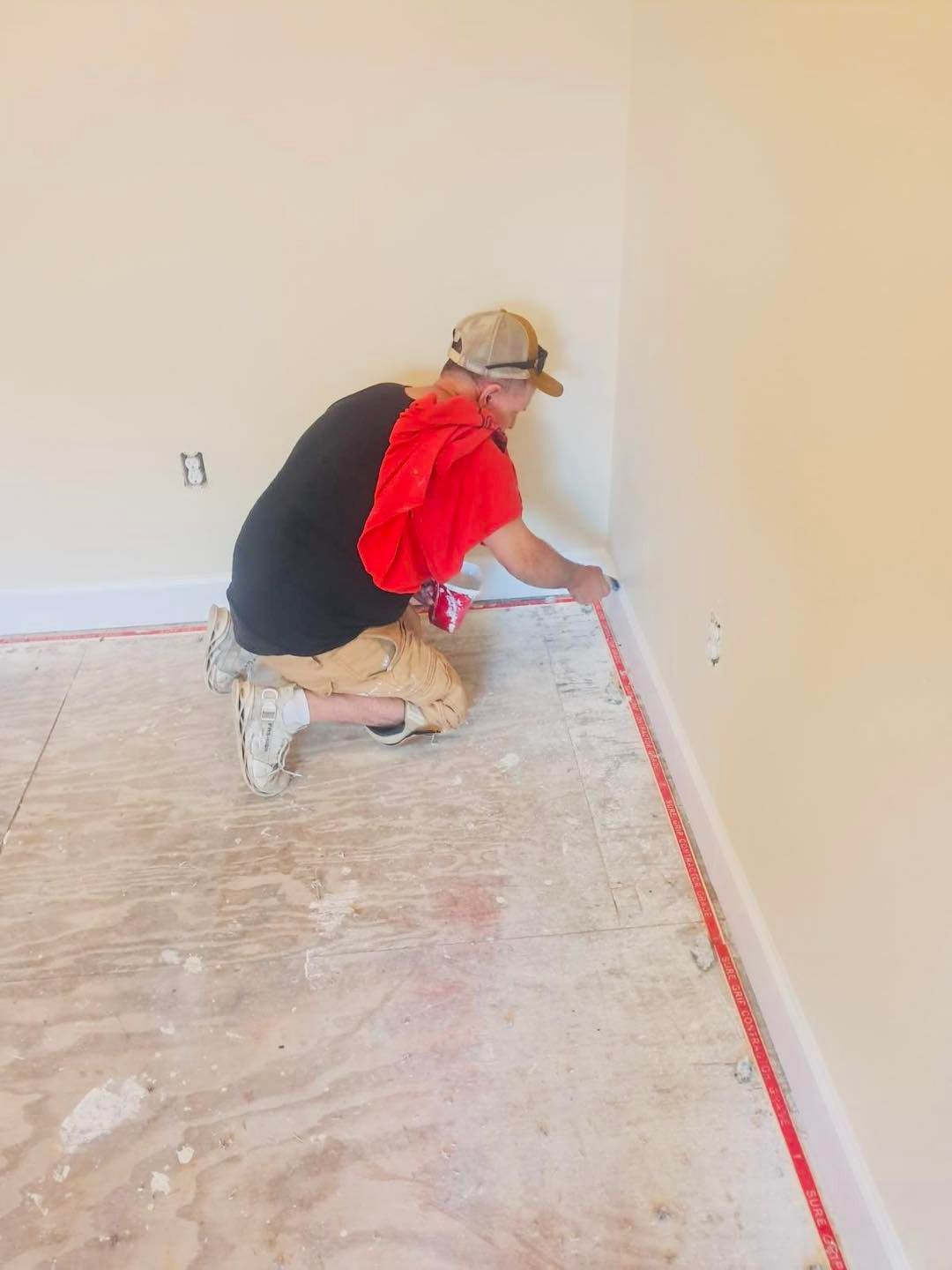 Person kneeling, measuring a wall corner in a room under construction, red tape along baseboard.