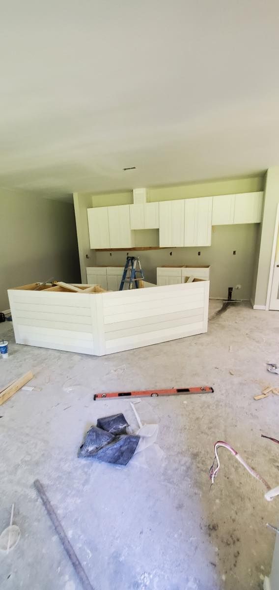 Interior construction site, unfinished kitchen island and cabinets, concrete floor, ladder in the background.