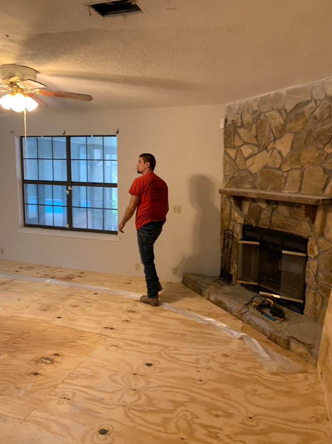 Man in red shirt surveys a room with plywood flooring, window, and stone fireplace.