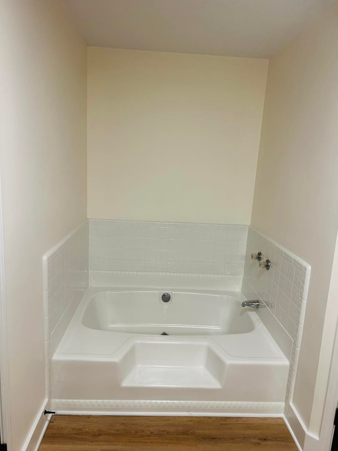 A white built-in bathtub with tiled backsplash and two steps, set in a room with light beige walls.