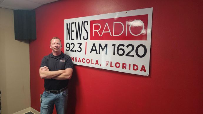 Man stands with arms crossed in front of a News Radio sign on a red wall in Pensacola, Florida.