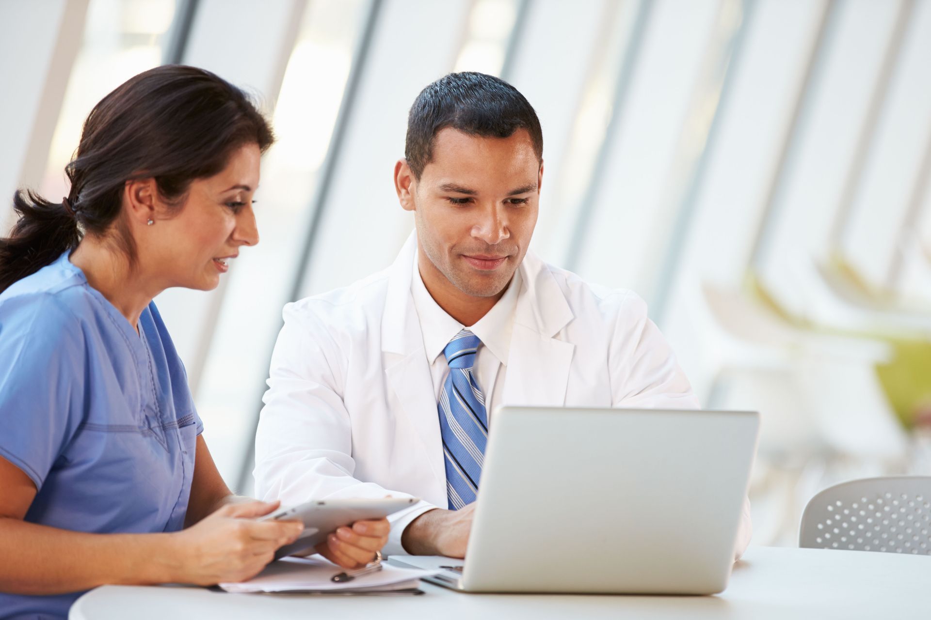 A nurse and a doctor in a white coat looking at a laptop screen together while sitting at a table in a bright office.