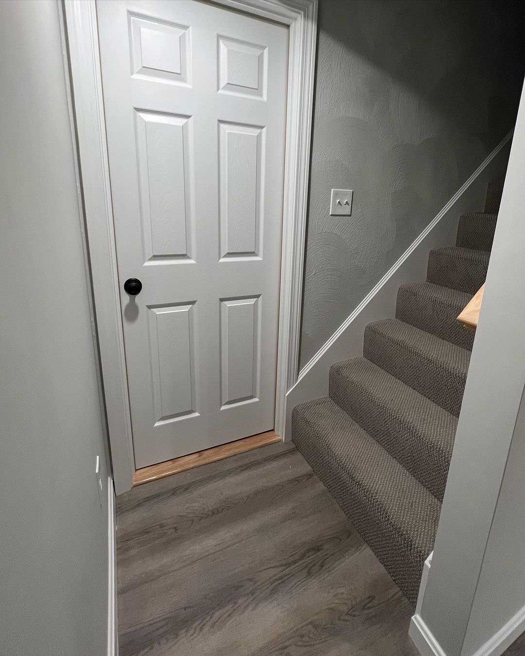 A white door and carpeted stairs in a hallway with gray walls and wooden flooring.