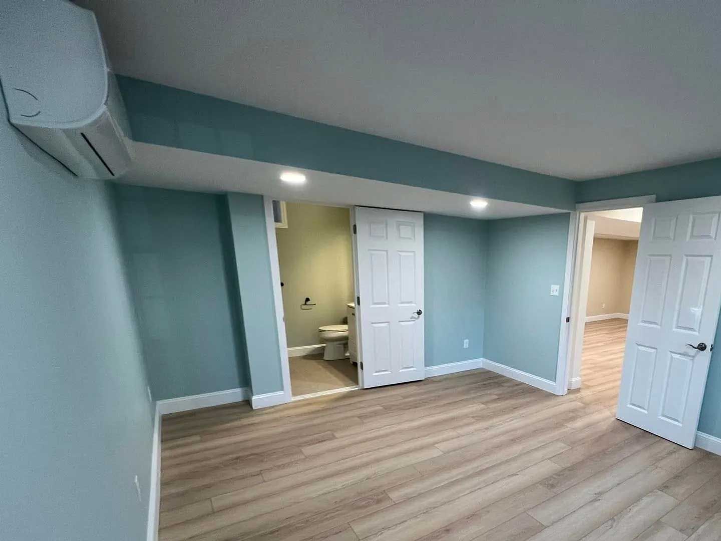 A room with teal walls, light wood floors, and white doors leading to a bathroom and another room.