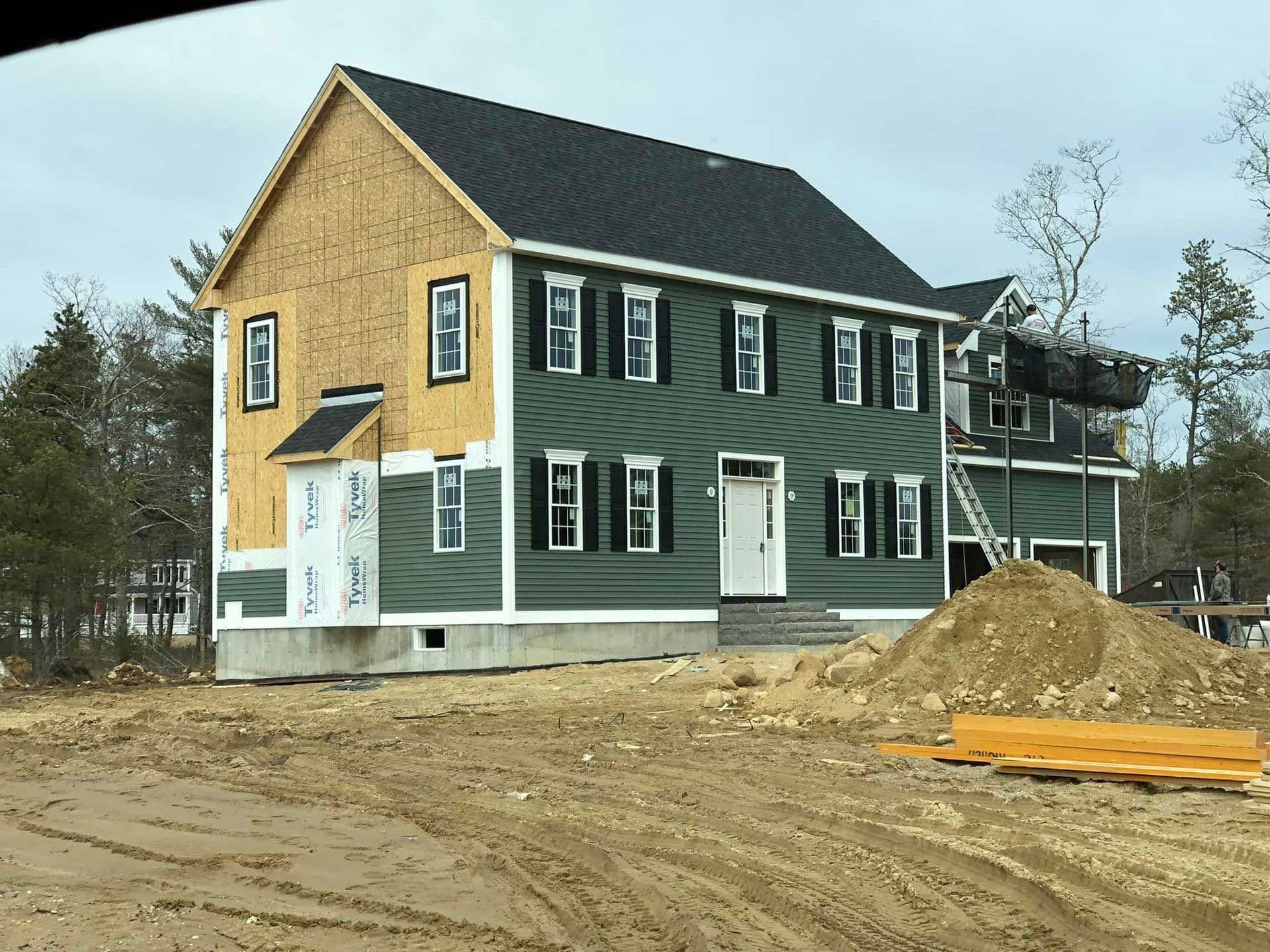 Two-story house under construction, green siding, black shutters, set on a brown dirt lot.
