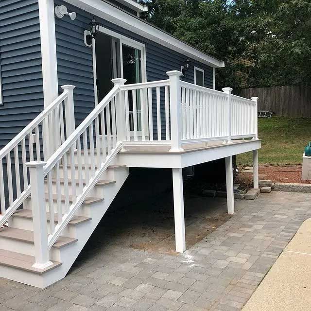 White deck with stairs attached to a blue-sided house, grey brick patio.