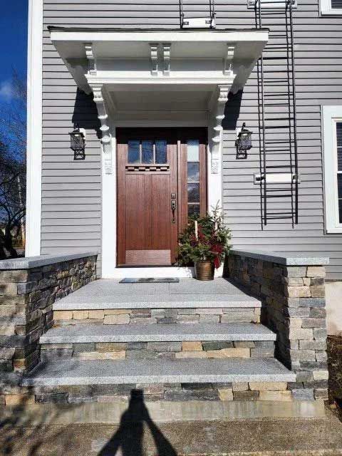 Gray house with brown door, stone steps, white awning. Christmas decorations are present.