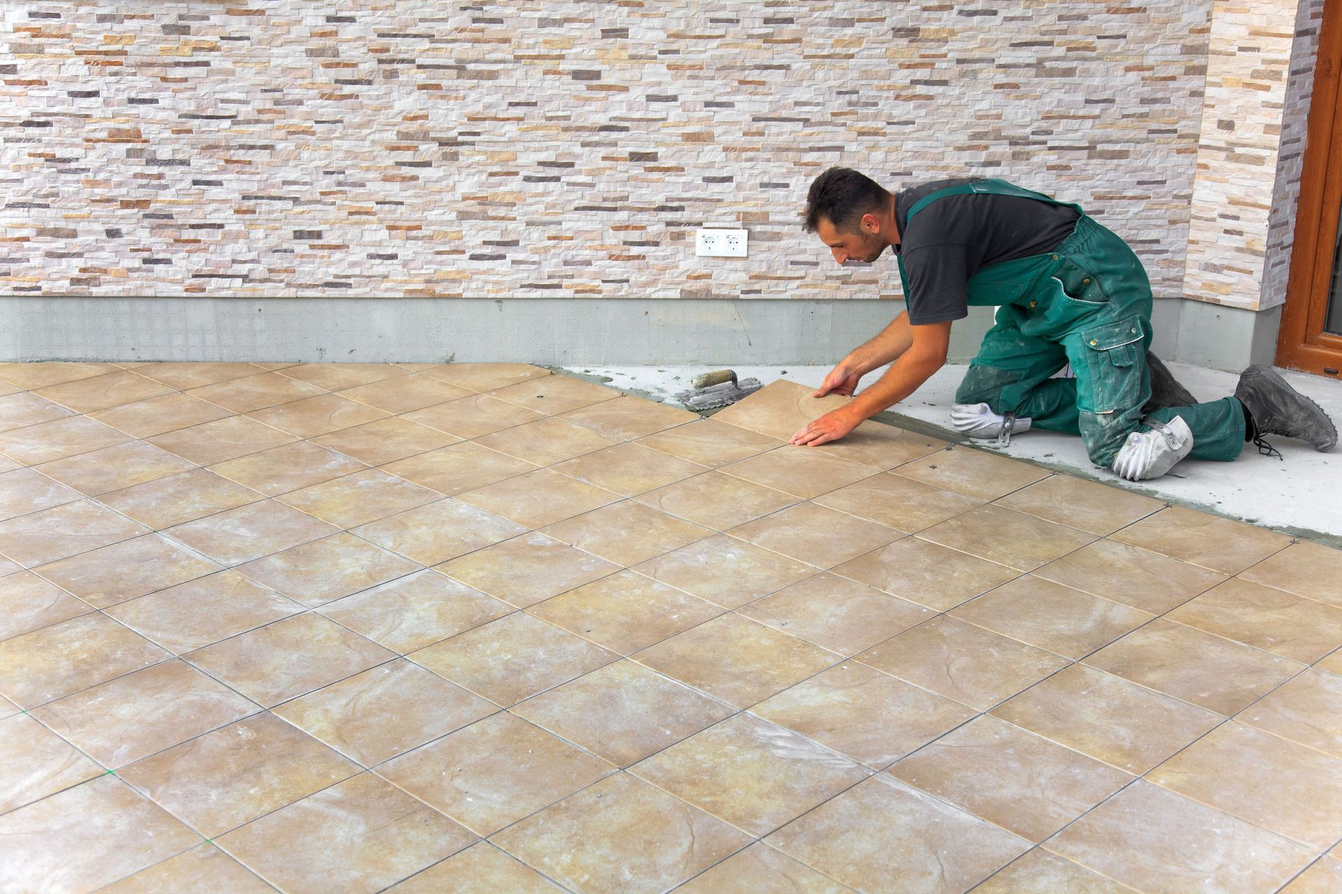 Man kneeling, installing brown tile on a patio next to a stone-textured wall.