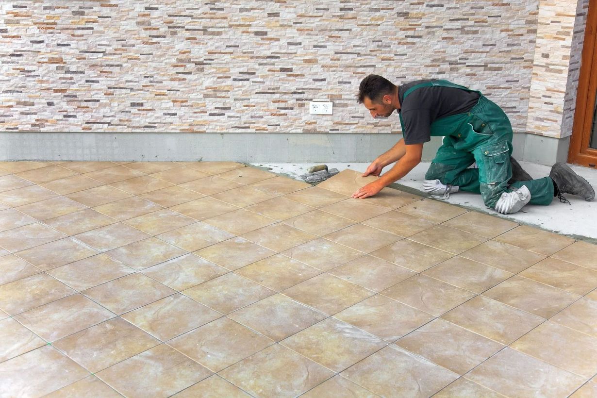 Man installing beige square tiles on a patio. He kneels, working with tiles near a stone-like wall.