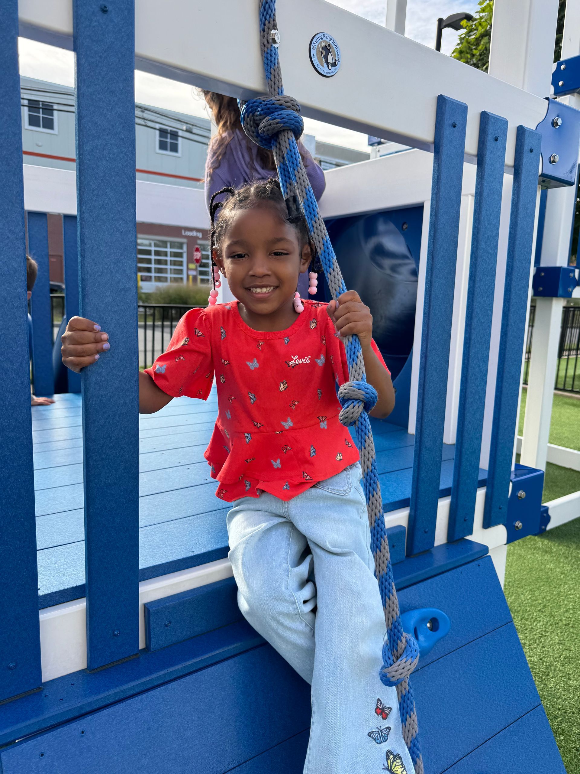 Girl with curly hair smiles, holding on to playground structure; wearing orange shirt, pink jacket, and black pants.