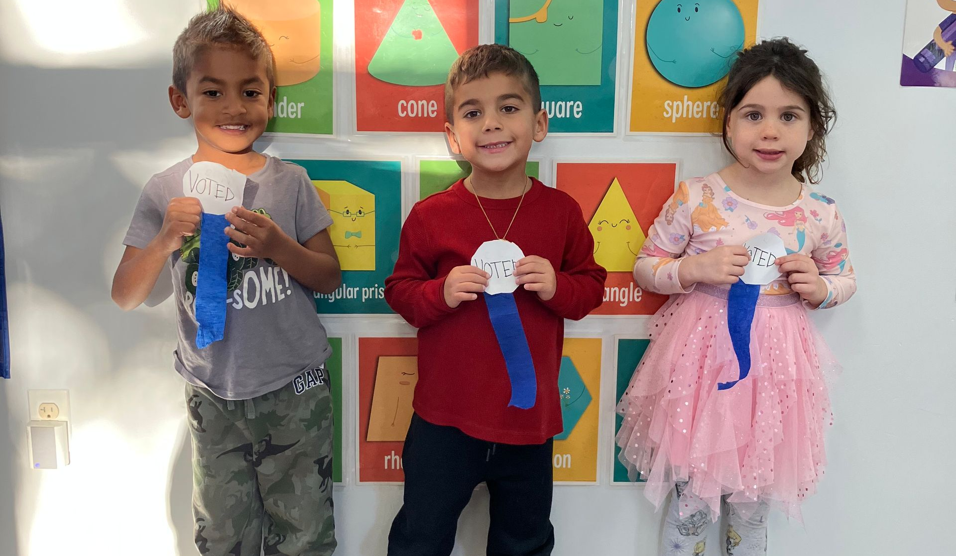 Two young children smiling and sitting next to each other outside. One in leopard jacket, the other in a light green coat.