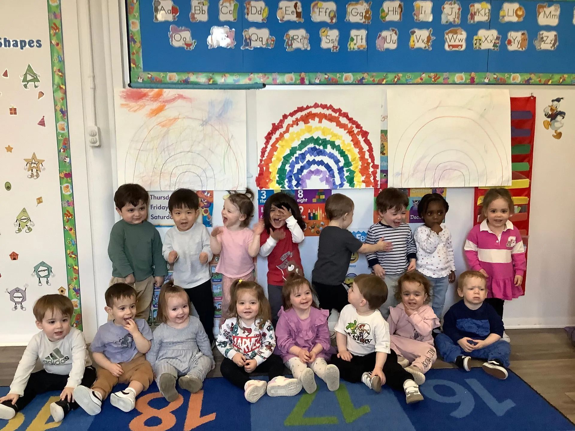 Children in a classroom pose with a rainbow art project.
