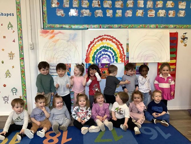 Children in a classroom pose with a rainbow art project.