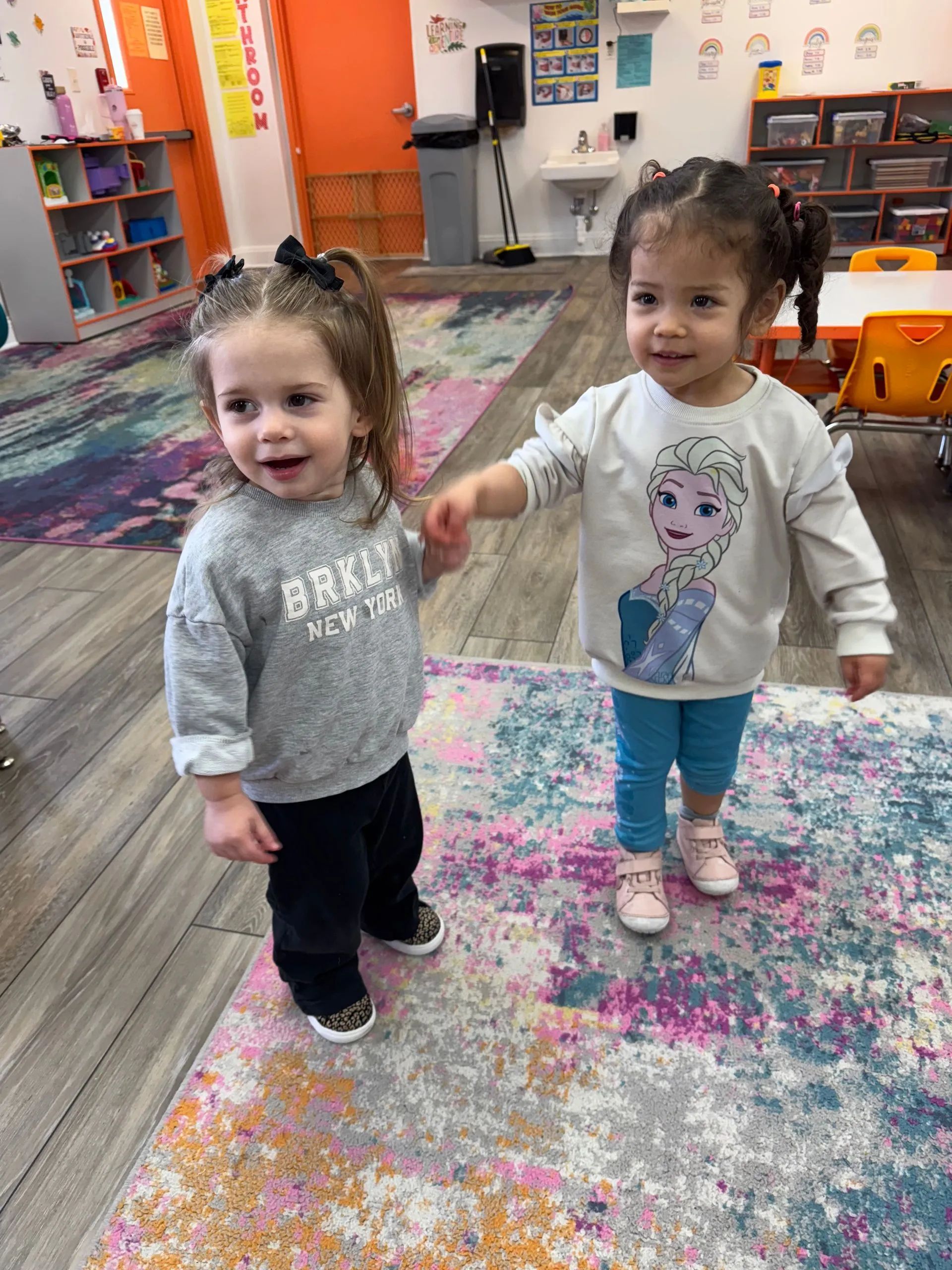 Two young girls holding hands, smiling in a colorful room with a rug. One has 