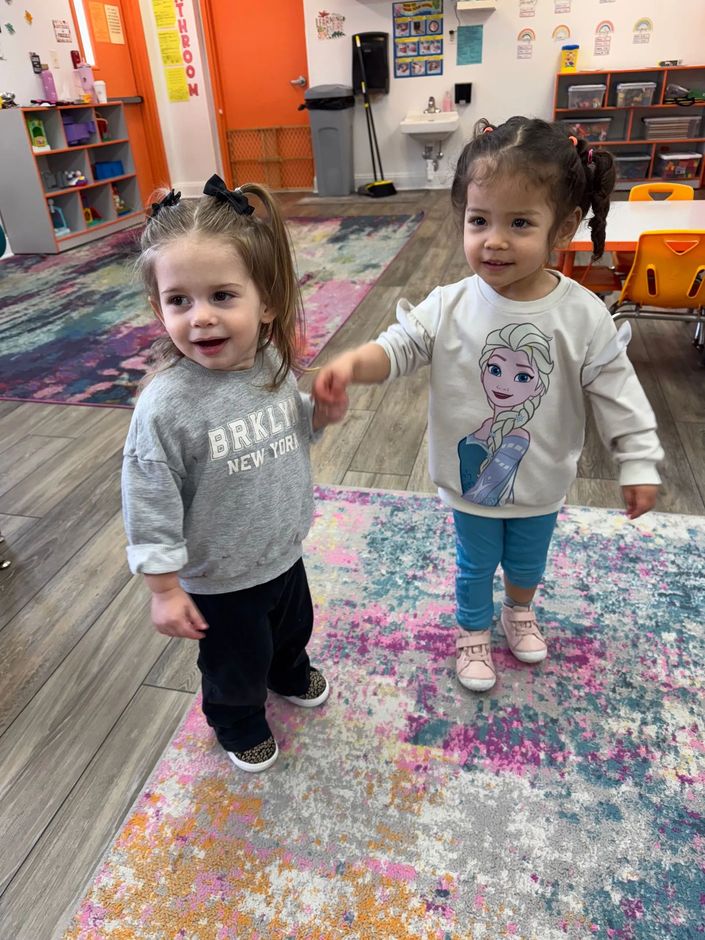 Two young girls holding hands, smiling in a colorful room with a rug. One has 