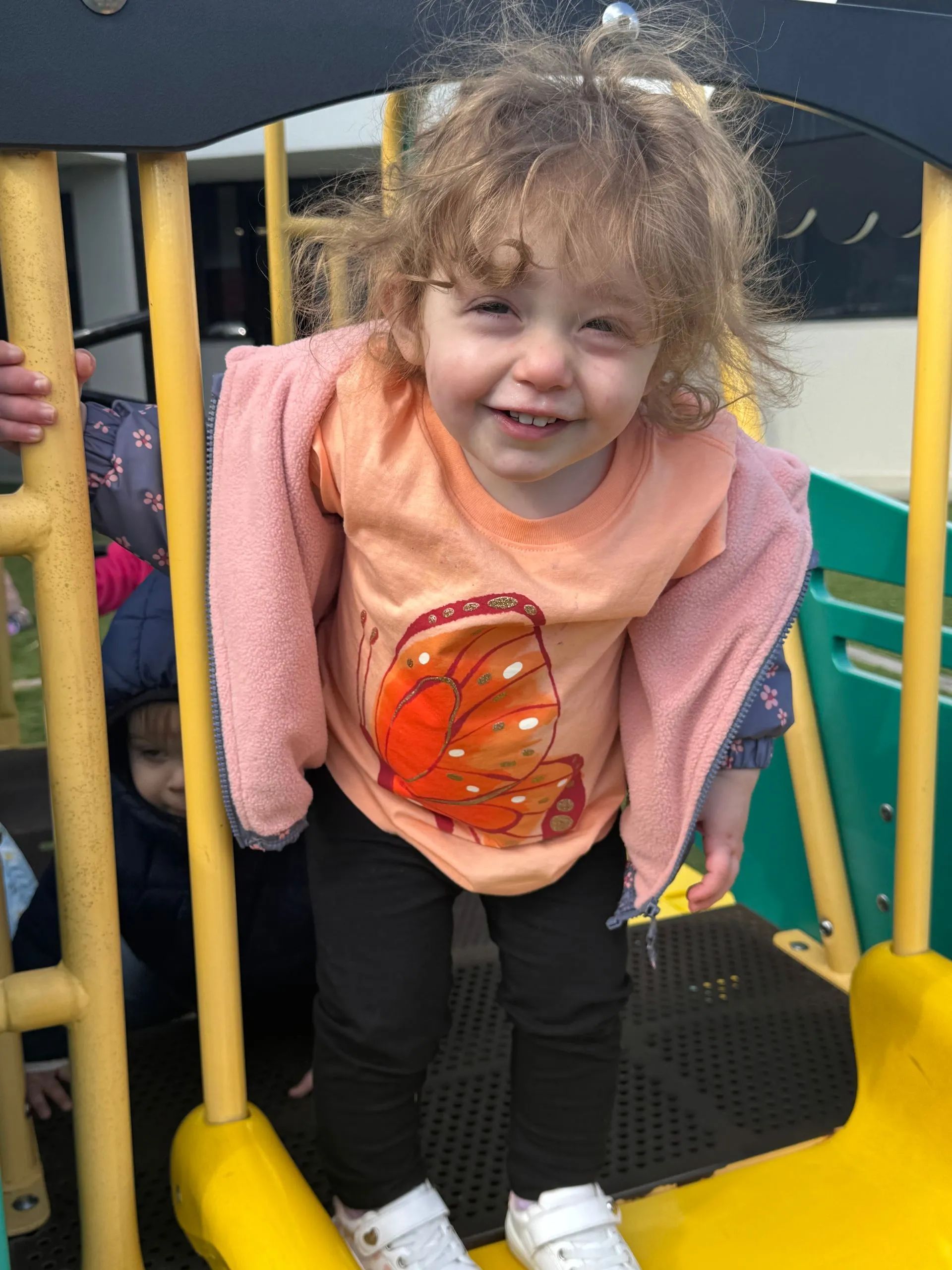 Girl with curly hair smiles, holding on to playground structure; wearing orange shirt, pink jacket, and black pants.